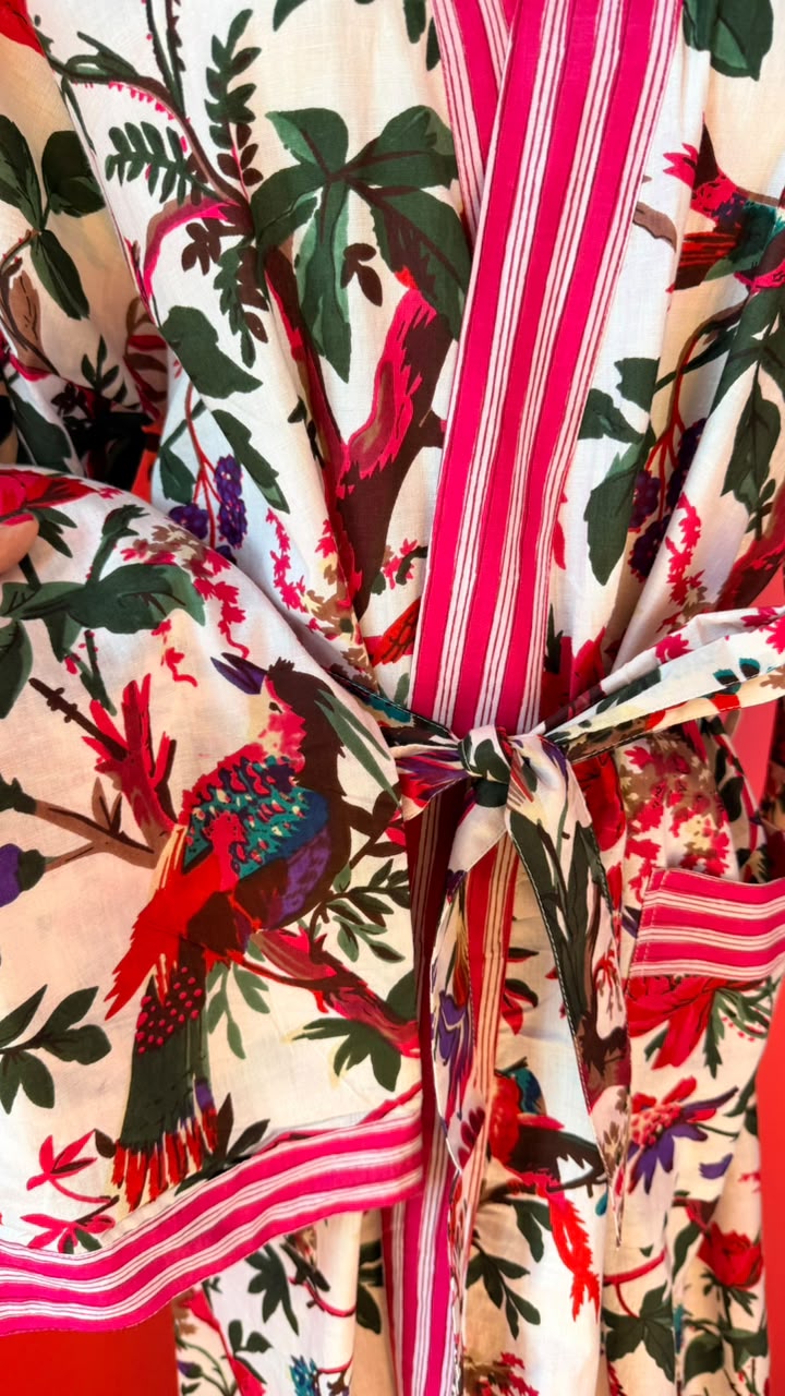A close-up of a patterned kimono robe with a tie belt, featuring a vibrant print of birds, foliage, and pink-and-white striped trim on a cream background.