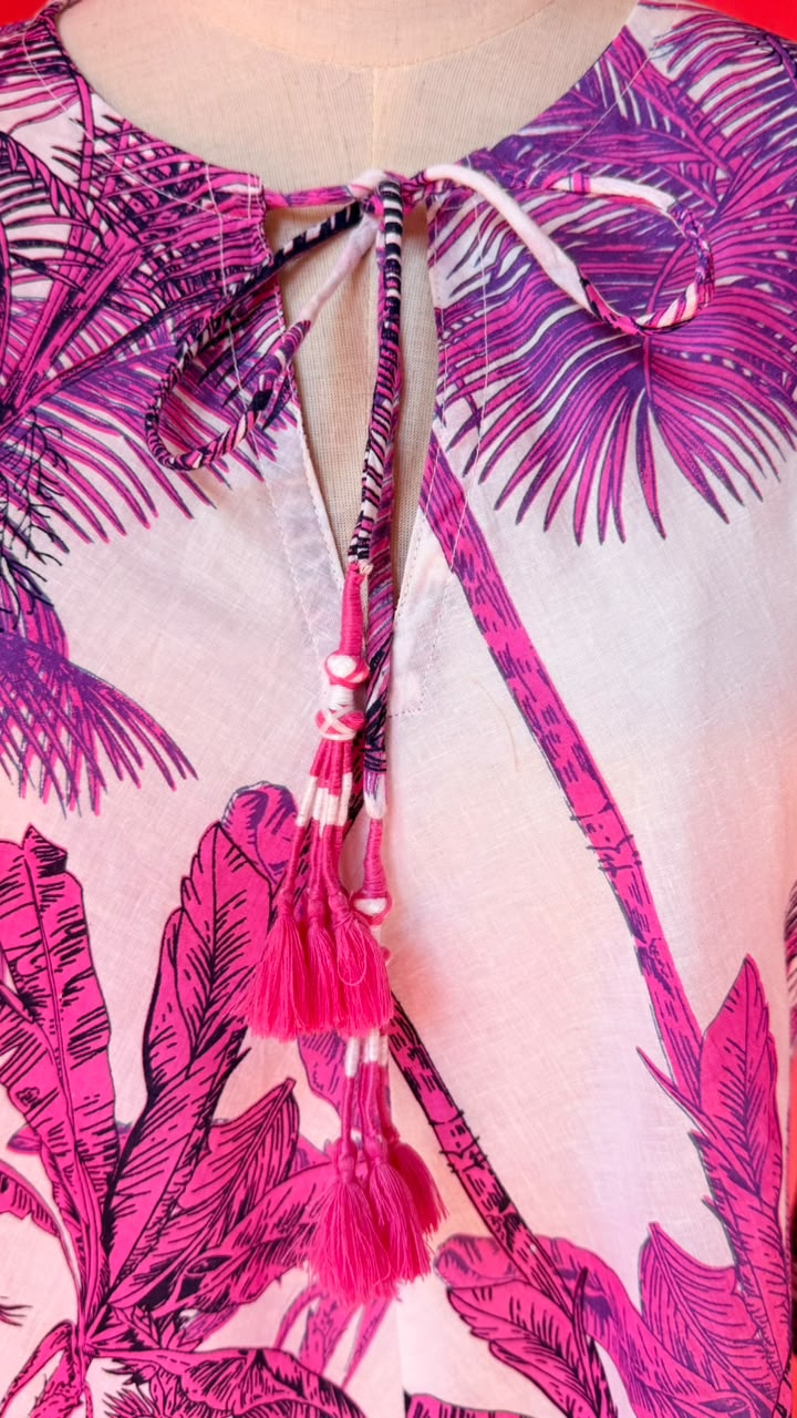 Close-up of a tropical print top featuring bright pink and magenta palm leaves on a pale pink background, with a tie-neck detail and tassel accents.