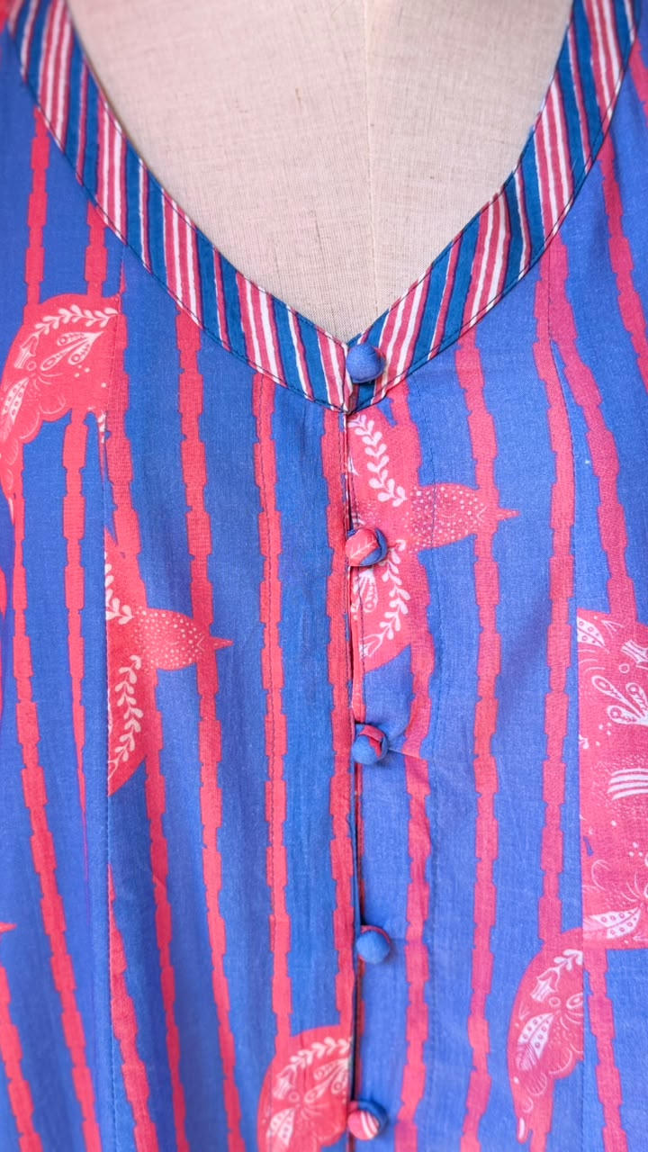 Close-up of a blue and pink striped fabric garment on a beige dress form, featuring vertical stripes, pink paisley motifs, and a striped V-neck collar.