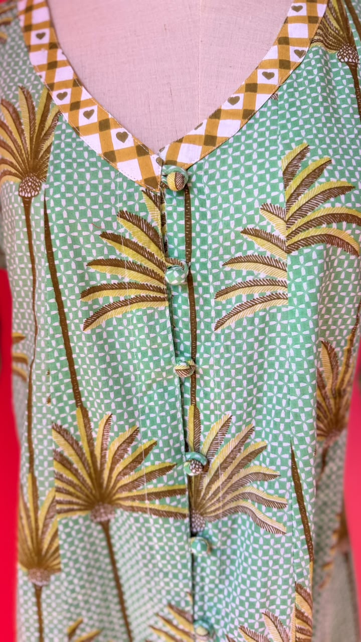 A close-up of a patterned tunic top on a pink dress form. The fabric features a mint green and white geometric checkered base with embroidered golden-brown palm tree motifs and a gingham trim in orange, white, and brown.