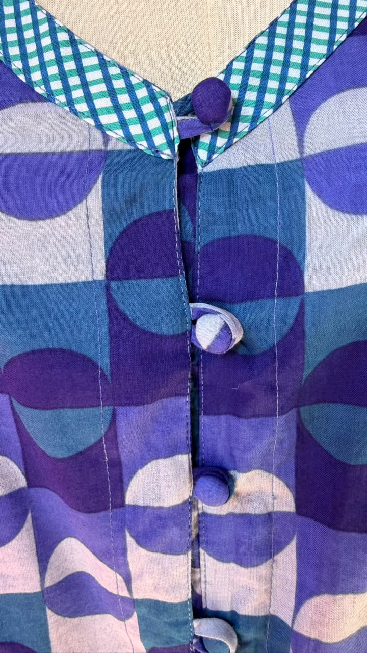 Close-up of a patterned blouse on a mannequin, featuring a geometric design in shades of purple, blue, and white, with a teal and white gingham collar and matching decorative buttons.