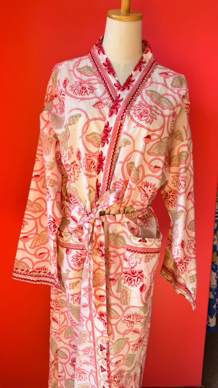 A long-sleeve patterned robe displayed on a mannequin torso against a bright red background. The robe has a cream base with pink, red, and beige paisley-style floral motifs and matching trim along the collar and cuffs.