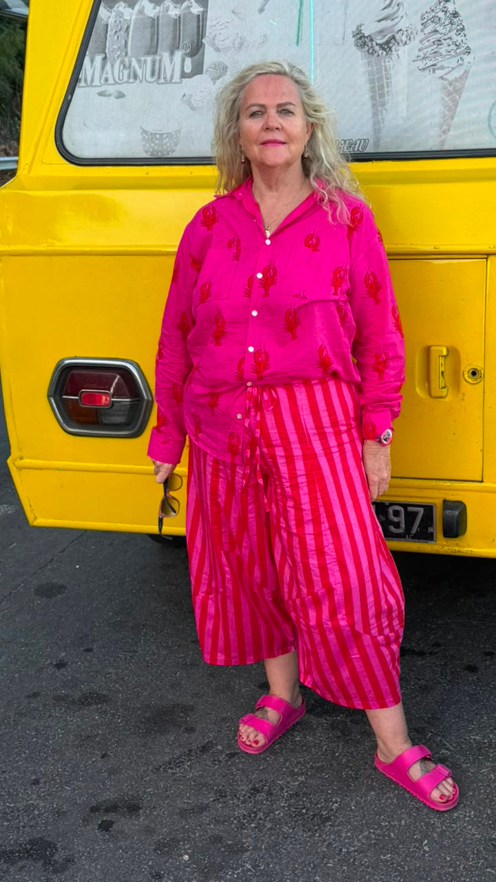 A woman stands in front of a bright yellow vehicle wearing a vibrant pink matching set consisting of a long-sleeve button-up shirt with red lobster embroidery and matching pink-and-white vertically striped cropped pants, paired with pink slide sandals.