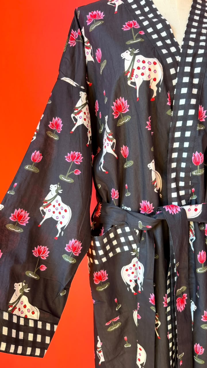 A black kimono robe featuring a whimsical print of white cows with red spots, pink lotus flowers, and green leaves, accented by a black and white checkered border on the collar, cuffs, and waist tie, set against a solid red background.