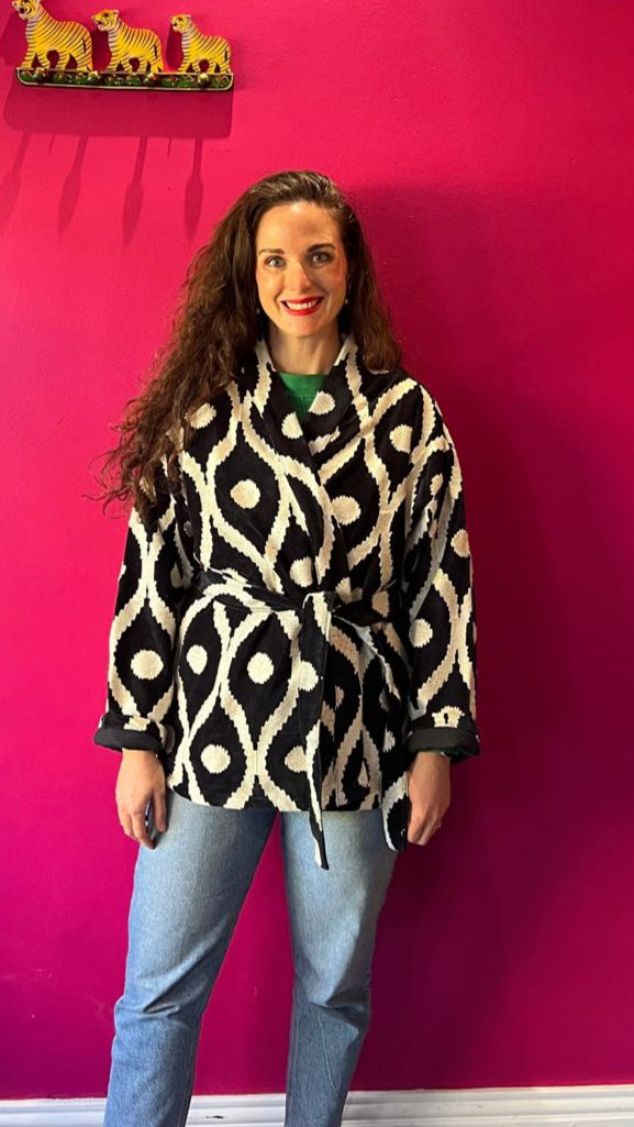 Woman wearing a patterned coat against a pink wall with three small yellow animals on a shelf. Gypsey rose