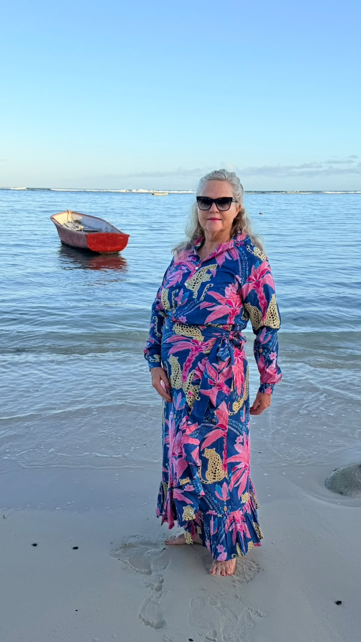 A woman wearing a vibrant blue, pink, and gold tropical floral maxi dress and sunglasses stands on a sandy beach with calm water and a small red boat in the background.
