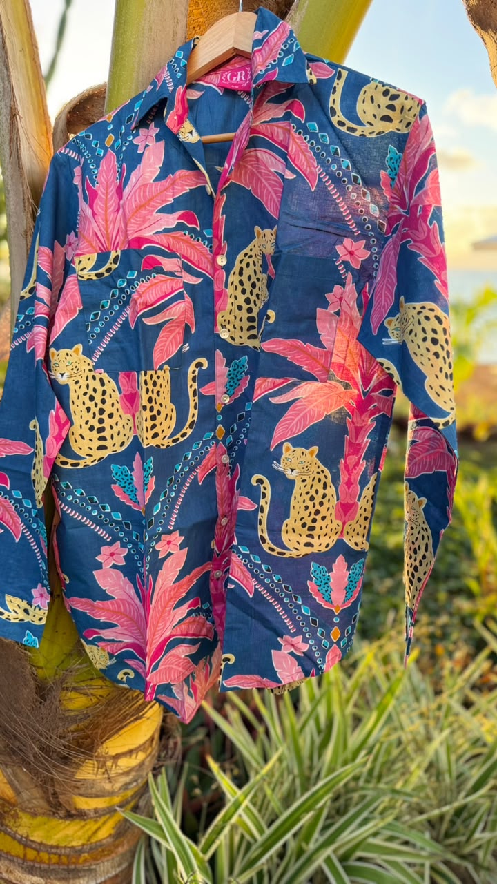 A long-sleeve button-up shirt with a vibrant tropical print featuring pink palm leaves, gold cheetahs, and teal patterns on a navy blue background, hanging on a wooden hanger outdoors against a palm tree trunk.