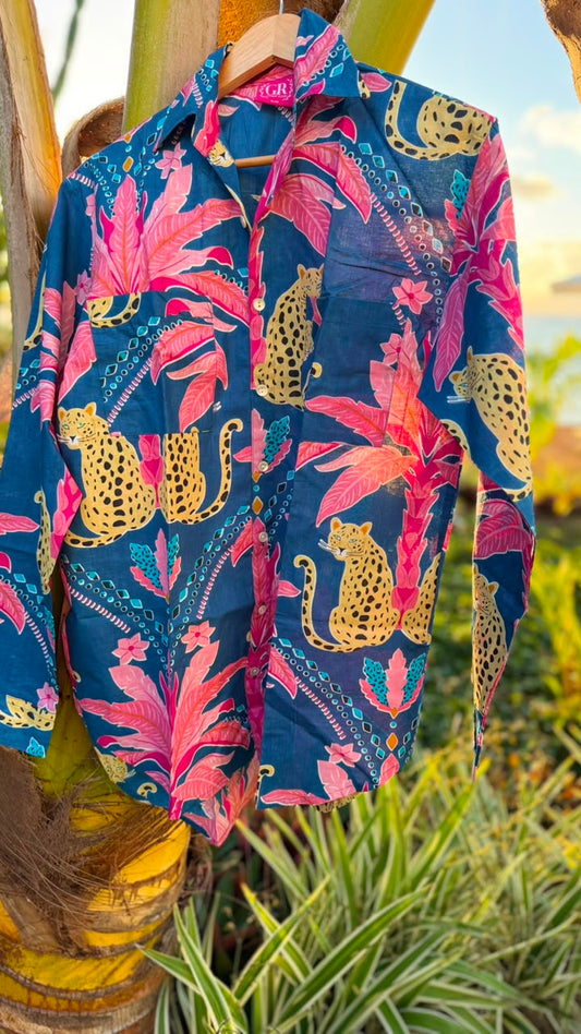 A long-sleeve button-up shirt with a vibrant tropical print featuring pink palm leaves, gold cheetahs, and teal patterns on a navy blue background, hanging on a wooden hanger outdoors against a palm tree trunk.