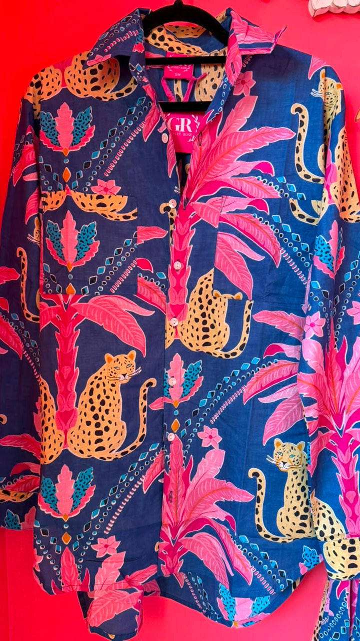 A long-sleeve button-up shirt hanging on a black hanger against a bright red wall. The shirt features a bold tropical print with pink palm leaves, yellow jaguars, and blue geometric patterns on a navy base.