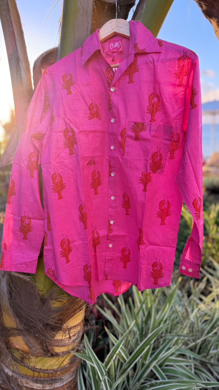 A bright pink long-sleeve button-up shirt with red lobster motifs, hanging on a wooden hanger outdoors against a palm tree trunk.