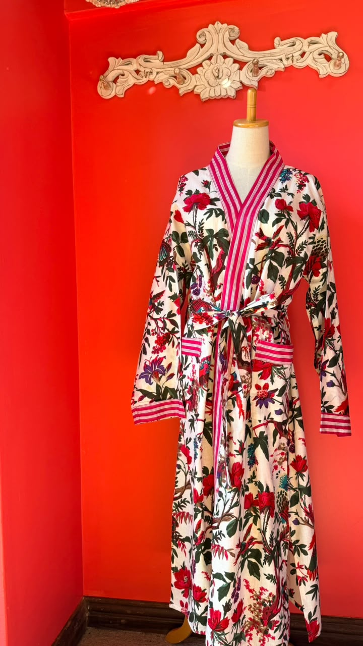 A long-sleeve floral robe displayed on a mannequin torso against a bright red wall. The robe has a cream base with red, pink, green, and black botanical print, plus pink striped trim and a matching tie belt.