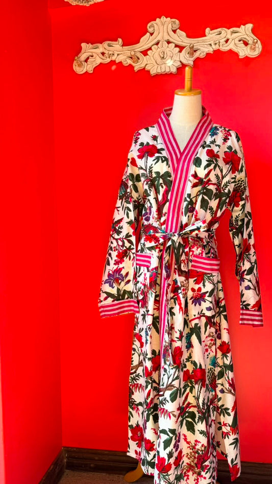 A long-sleeve floral robe displayed on a mannequin torso against a bright red wall. The robe has a cream base with red, pink, green, and black botanical print, plus pink striped trim and a matching tie belt.