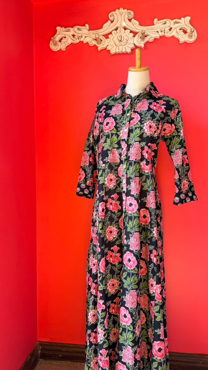 A long-sleeve button-up shirt dress with a vibrant pink, red, and green floral pattern on a black background, displayed on a mannequin against a bright red wall.