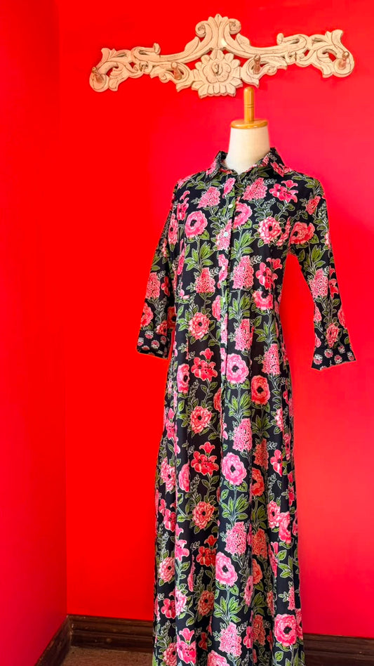 A long-sleeve button-up shirt dress with a vibrant pink, red, and green floral pattern on a black background, displayed on a mannequin against a bright red wall.