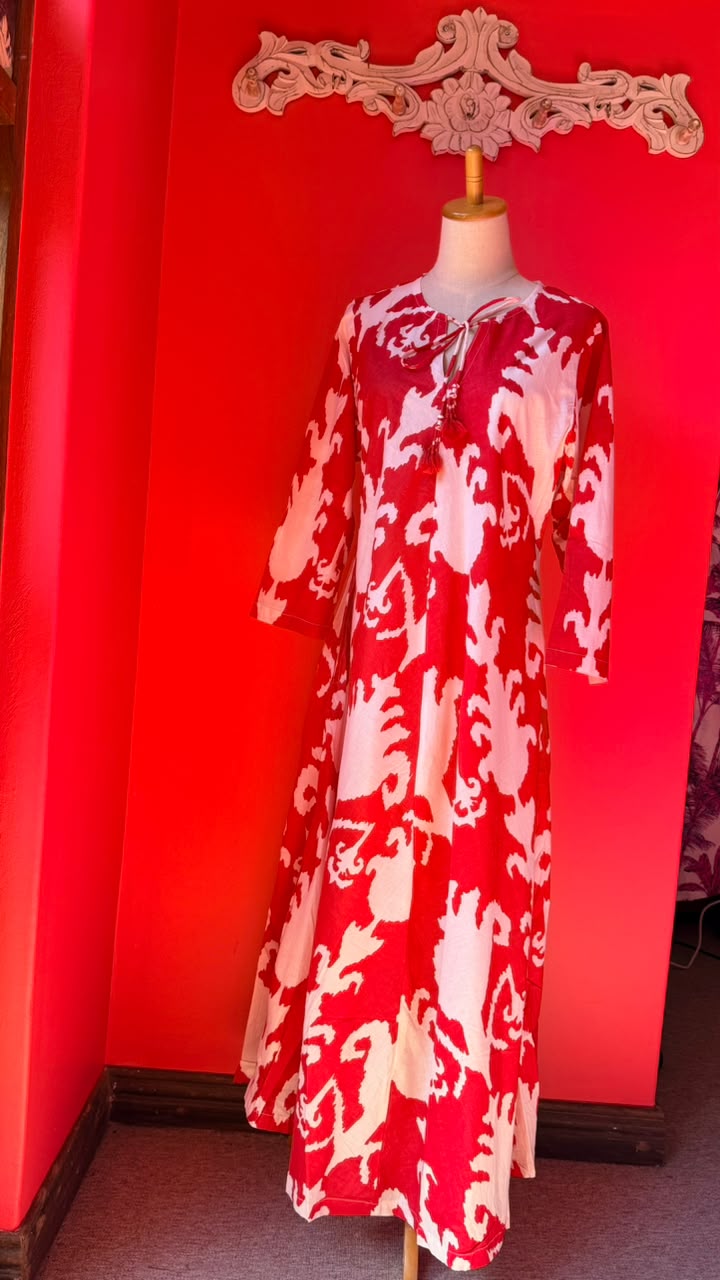 A red and white patterned maxi dress displayed on a mannequin in a room with a bright red wall and a decorative white wall piece above it.