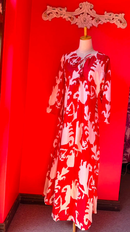 A red and white patterned maxi dress displayed on a mannequin in a room with a bright red wall and a decorative white wall piece above it.