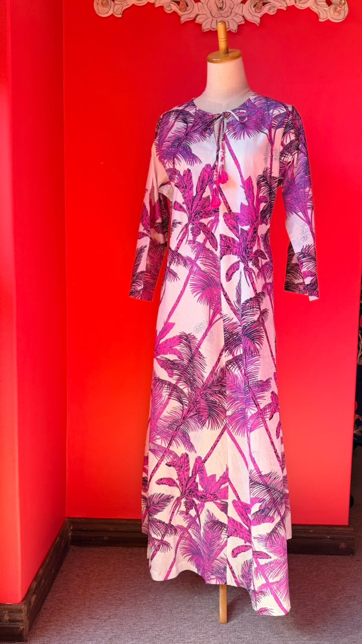 A long-sleeve maxi dress with a pink and purple palm tree print on a white base, displayed on a mannequin against a bright red wall.