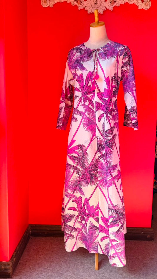 A long-sleeve maxi dress with a pink and purple palm tree print on a white base, displayed on a mannequin against a bright red wall.