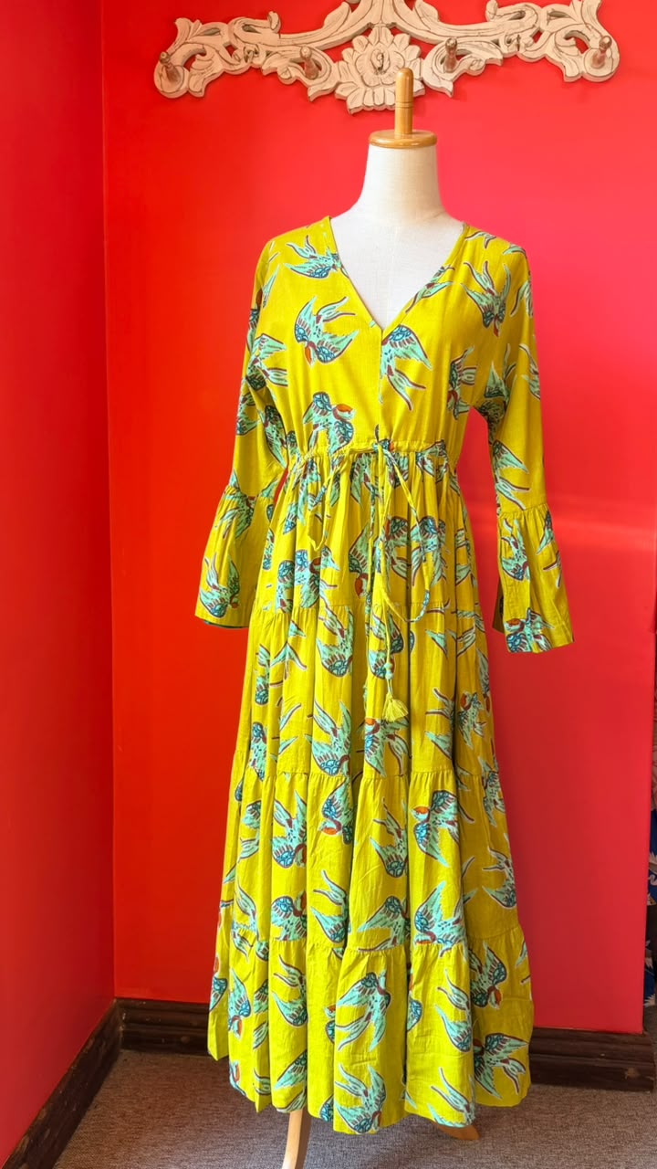 A yellow maxi dress with a blue and green tropical leaf print, displayed on a mannequin against a bright red wall.
