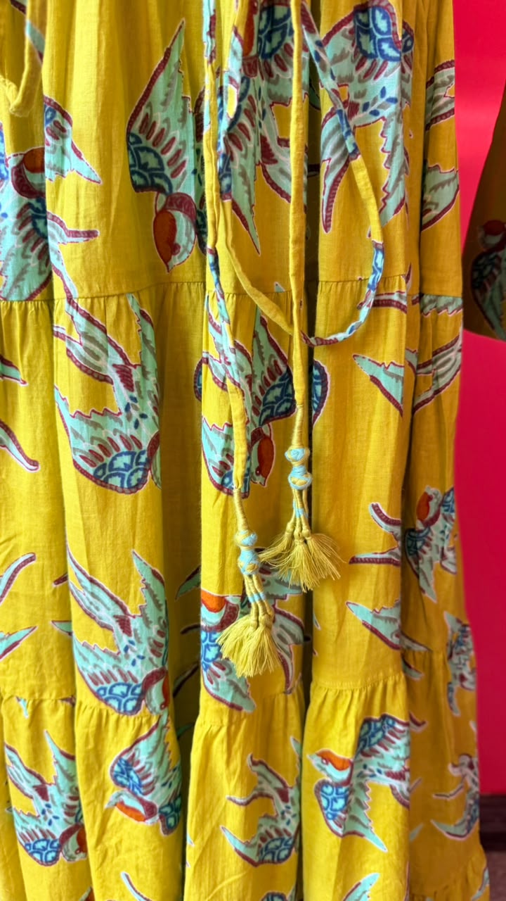 A close-up of a tiered yellow dress or long skirt with a colorful bird print in teal, red, and white, featuring a drawstring waist with tassels.