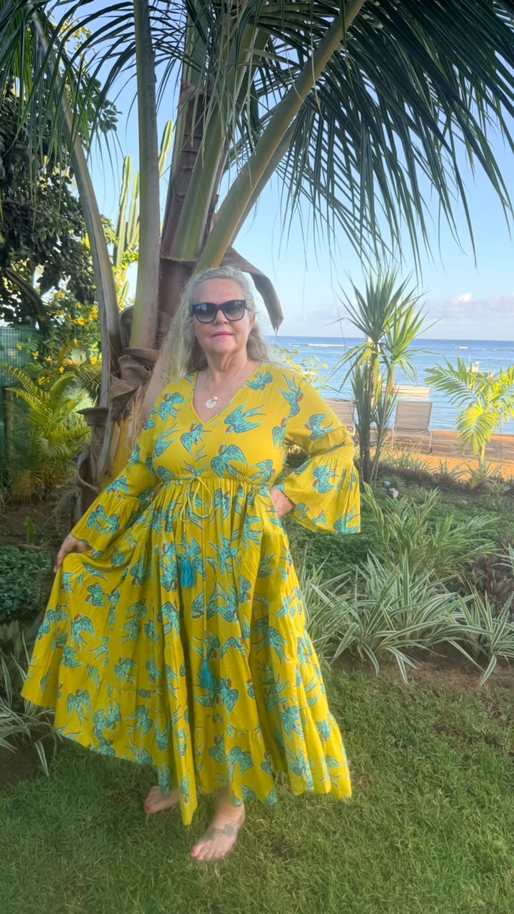 A woman wearing a yellow and teal tropical print maxi dress stands barefoot outdoors on a grassy lawn, with palm trees, a beach, and ocean visible in the background.