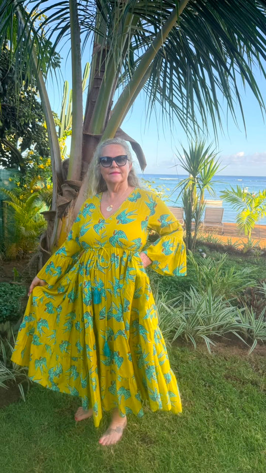 A woman wearing a yellow and teal tropical print maxi dress stands barefoot outdoors on a grassy lawn, with palm trees, a beach, and ocean visible in the background.
