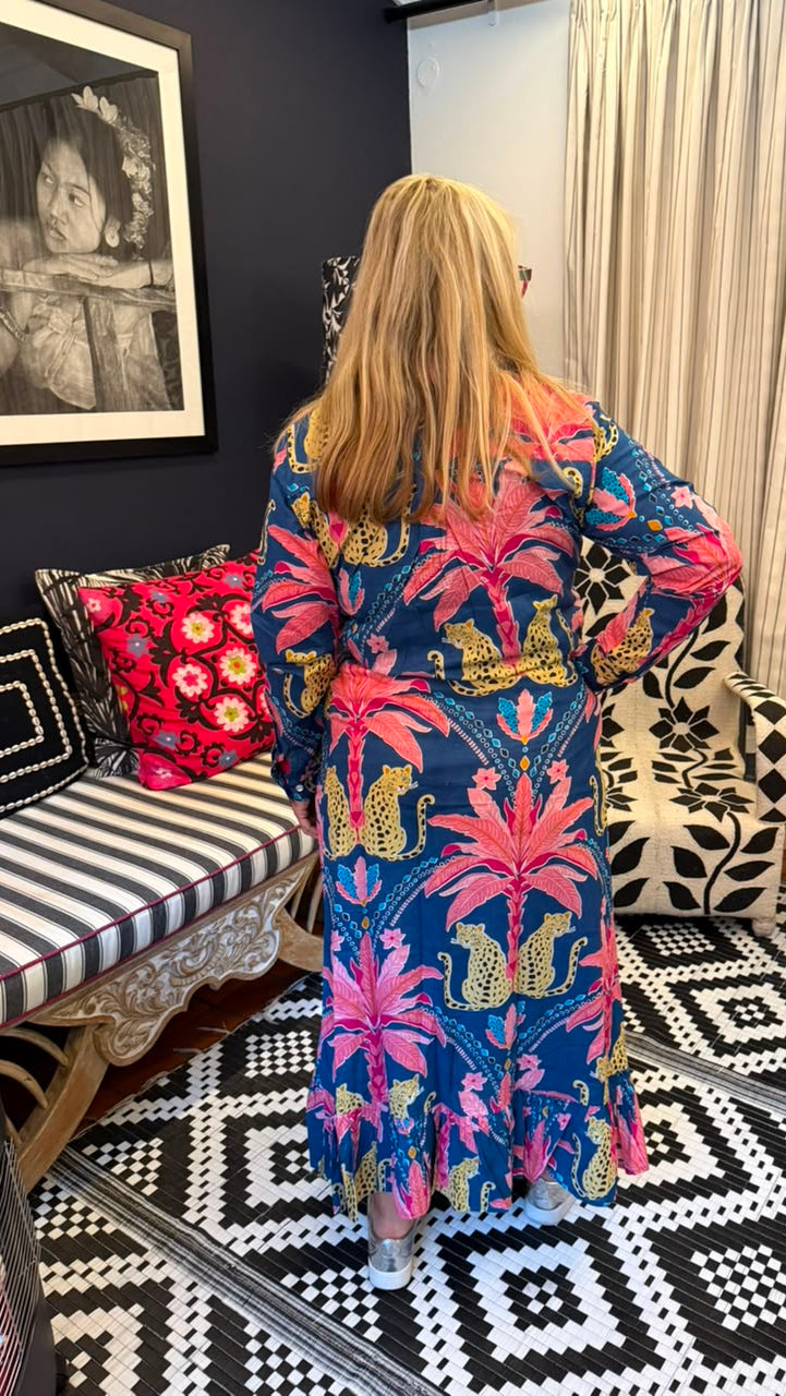 A woman stands with her back to the camera wearing a long-sleeve maxi dress with a vibrant tropical print. The dress features a navy blue base with large pink palm trees, yellow and orange birds, and green leaves.