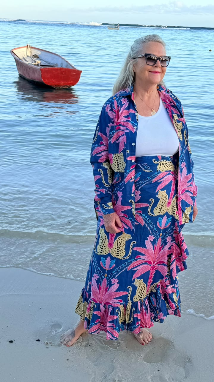 A woman stands on a sandy beach wearing a long blue maxi dress with a bold pink and gold tropical floral print. She is also wearing a white tank top and sunglasses.