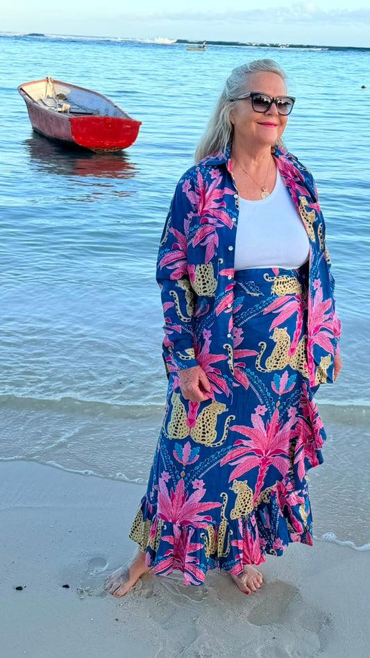 A woman stands on a sandy beach wearing a long blue maxi dress with a bold pink and gold tropical floral print. She is also wearing a white tank top and sunglasses.