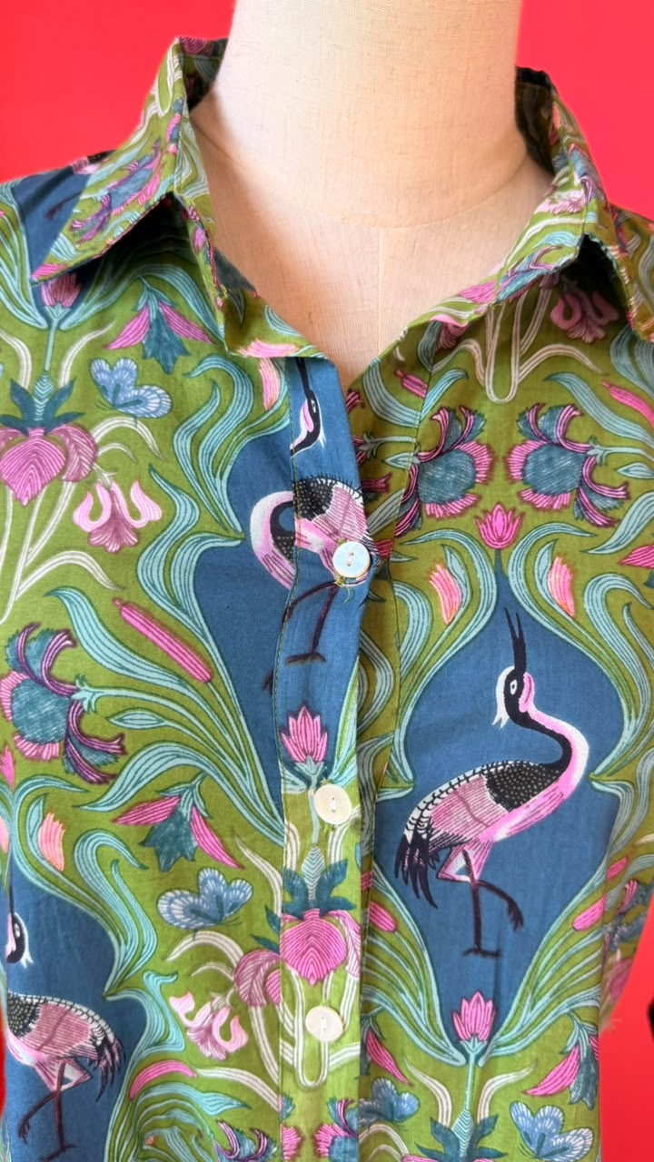A close-up of a button-front collared shirt displayed on a mannequin torso, featuring a bold botanical and flamingo print in green, teal, pink, and blue against a bright red background.