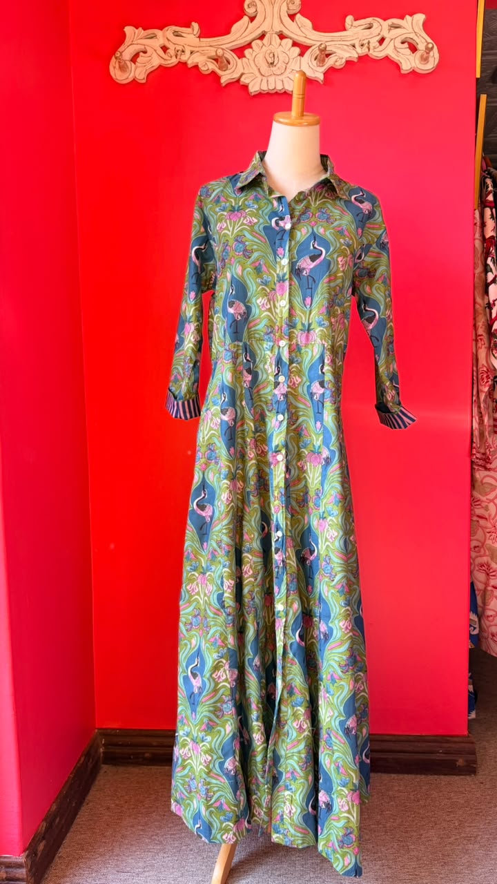 A long-sleeve button-front maxi dress with a vibrant green, blue, and pink flamingo print, displayed on a mannequin against a bright red wall.