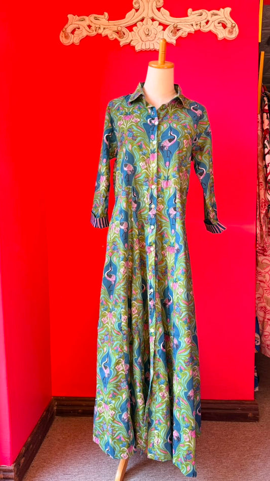 A long-sleeve button-front maxi dress with a vibrant green, blue, and pink flamingo print, displayed on a mannequin against a bright red wall.