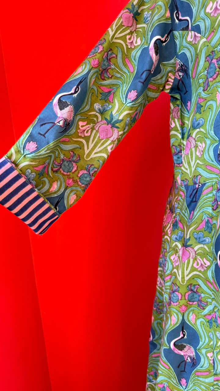 A close-up of a colorful patterned garment featuring blue flamingos, pink lotus flowers, and green and blue abstract swirls, with a striped cuff in navy and pink, set against a solid bright red background.