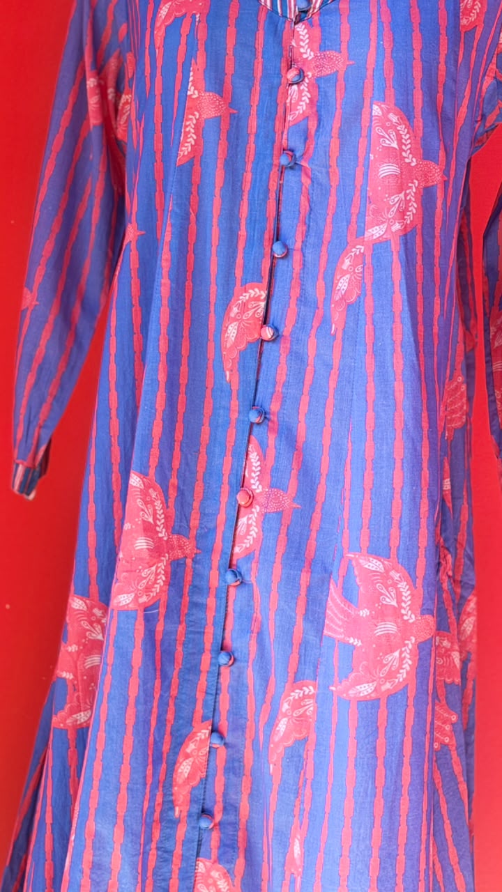 A close-up of a blue and coral patterned long-sleeve tunic with vertical red stripes and red floral motifs, featuring a buttoned front closure, set against a solid red background.