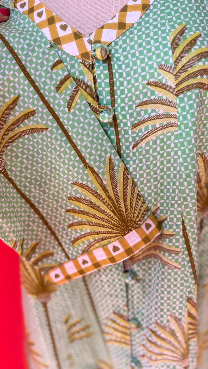 A close-up of a patterned garment featuring a mint green and white checkered base with brown and gold palm leaf motifs and orange plaid trim.