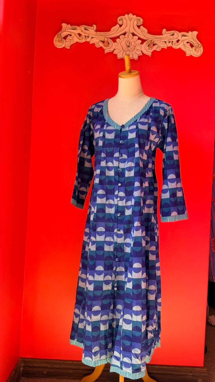 A long-sleeve button-front tunic dress displayed on a mannequin torso against a vivid red wall. The garment has an allover geometric arch pattern in shades of blue, purple, and white, with a checked trim.