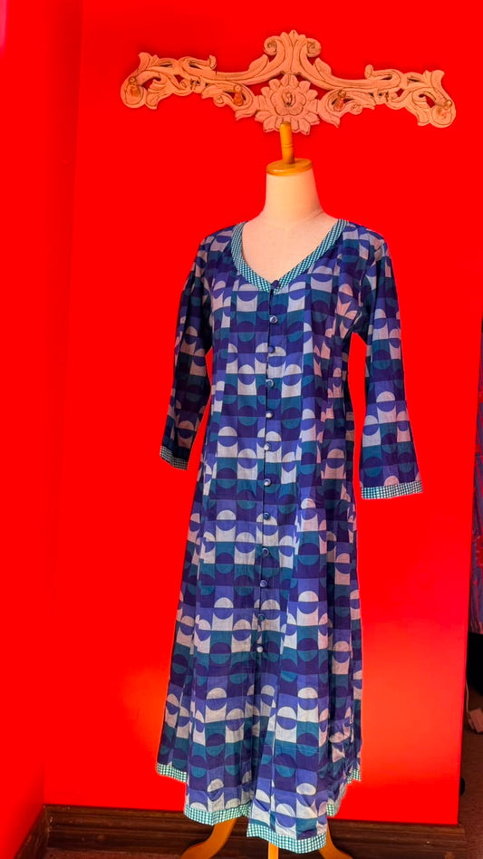 A long-sleeve button-front tunic dress displayed on a mannequin torso against a vivid red wall. The garment has an allover geometric arch pattern in shades of blue, purple, and white, with a checked trim.
