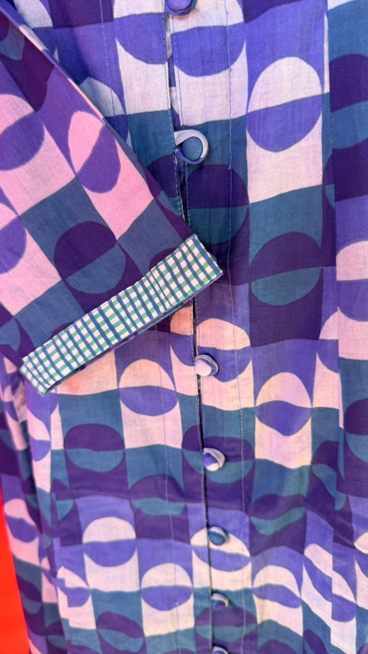 Close-up of a button-front garment with an abstract geometric print in purple, blue, pink, and teal, plus a gingham pocket flap.