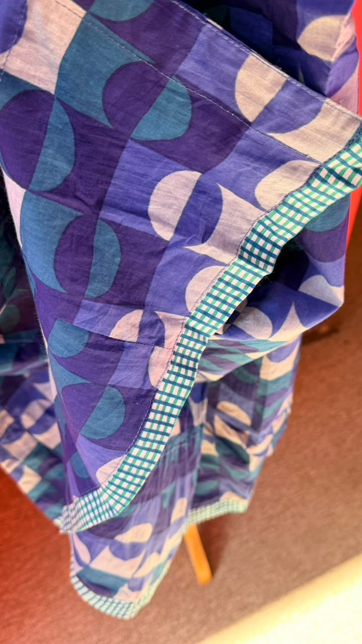 A close-up of a patterned fabric item, likely a scarf or wrap, featuring a geometric design in shades of purple, blue, and white, with a green and white gingham border.