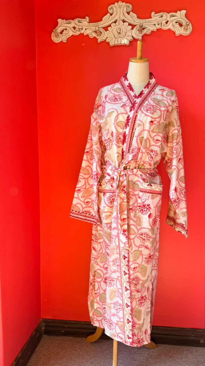 A long-sleeve patterned robe displayed on a mannequin against a bright red wall. The robe has a cream base with pink and red floral motifs and red trim.