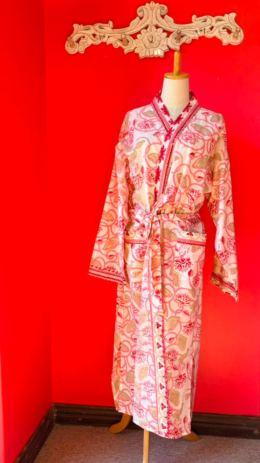 A long-sleeve patterned robe displayed on a mannequin against a bright red wall. The robe has a cream base with pink and red floral motifs and red trim.