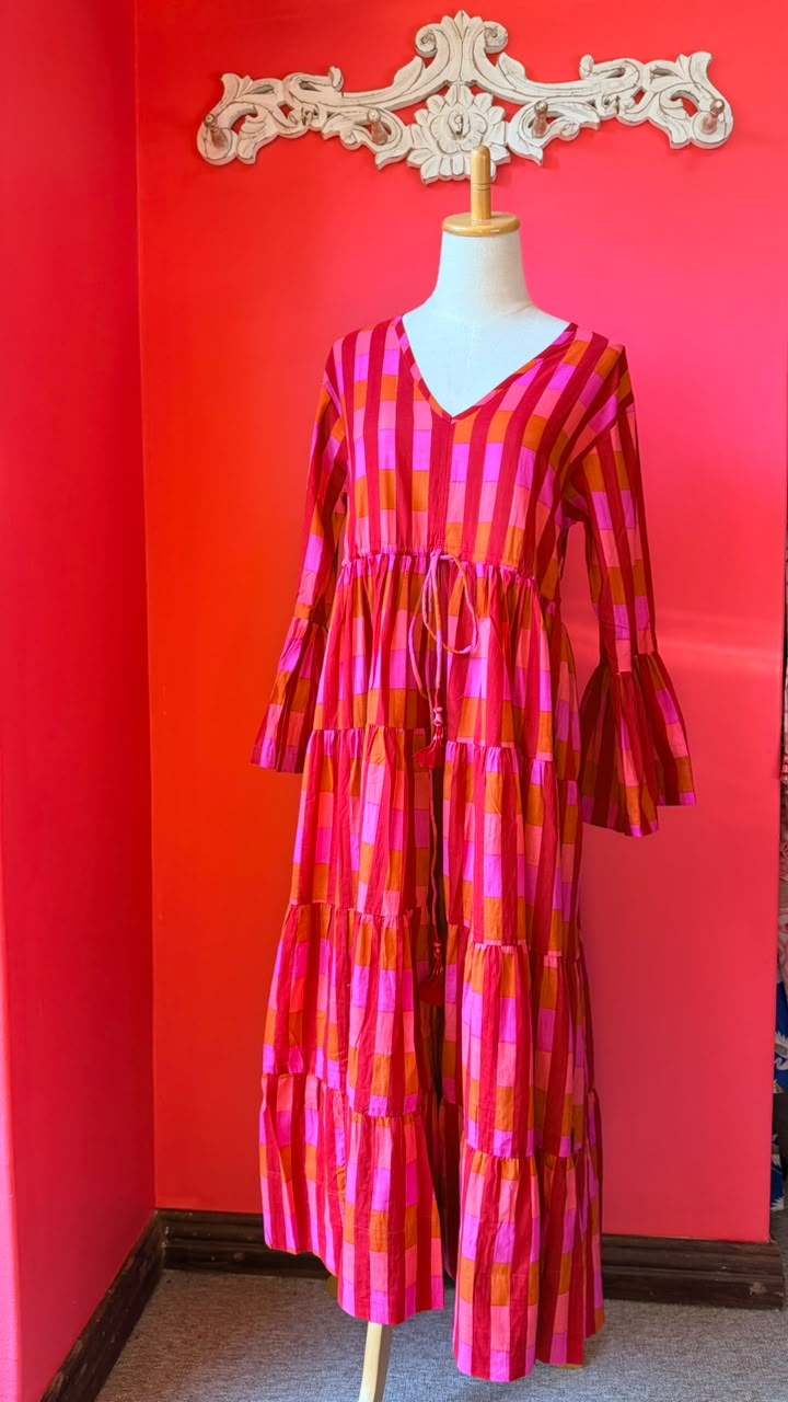 A tiered dress with a bold pink, orange, and red plaid pattern, displayed on a mannequin in front of a bright red wall.