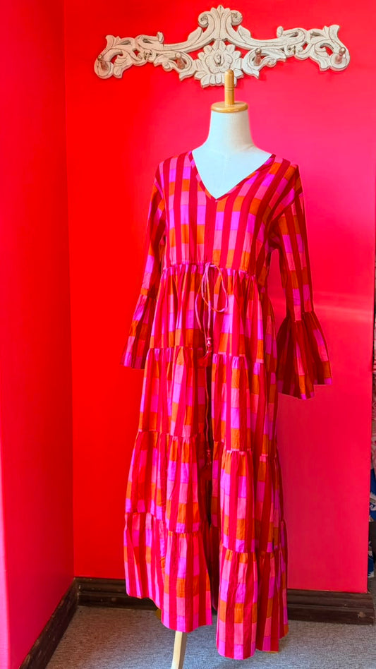 A tiered dress with a bold pink, orange, and red plaid pattern, displayed on a mannequin in front of a bright red wall.