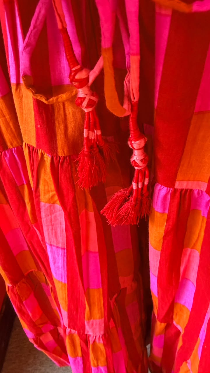 A close-up of a colorful patchwork fabric garment, likely a dress, featuring bright pink, red, orange, and light pink checkered patterns with tassel ties.