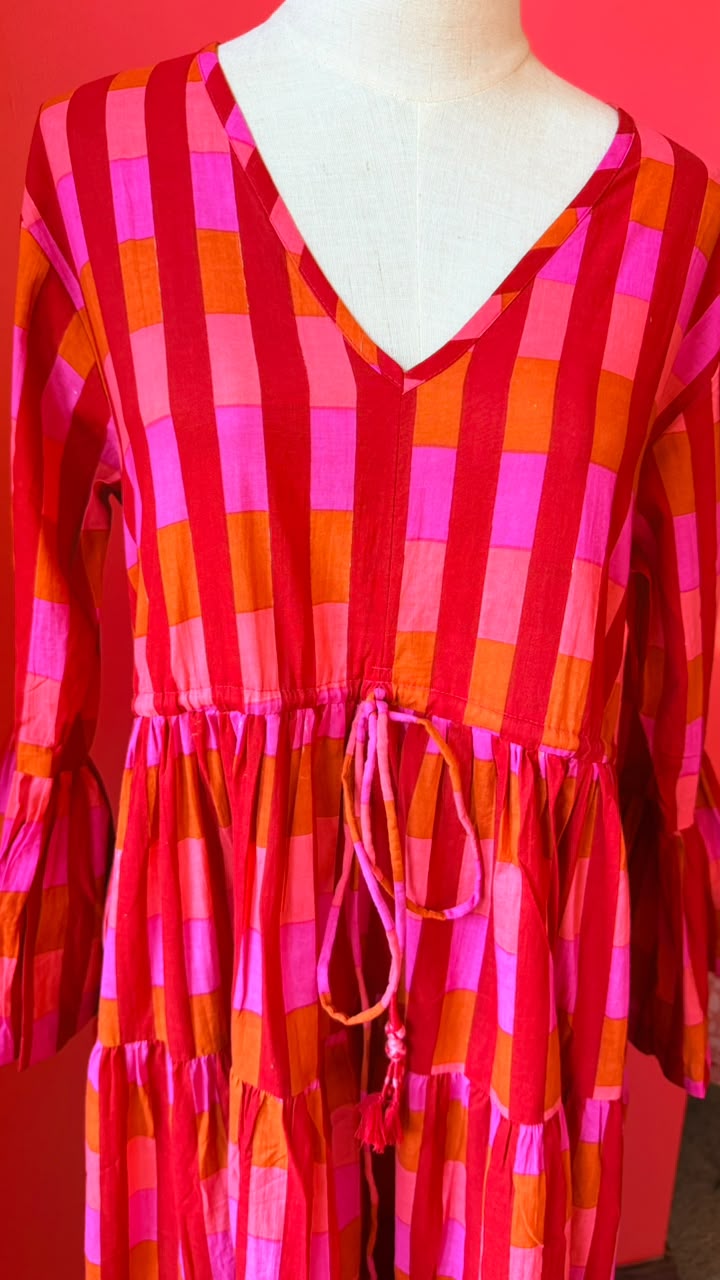 A tiered cotton dress with a bold red, orange, and pink checkered pattern, featuring a V-neckline, elbow-length sleeves, and a drawstring waist, displayed on a white dress form against a solid red background.