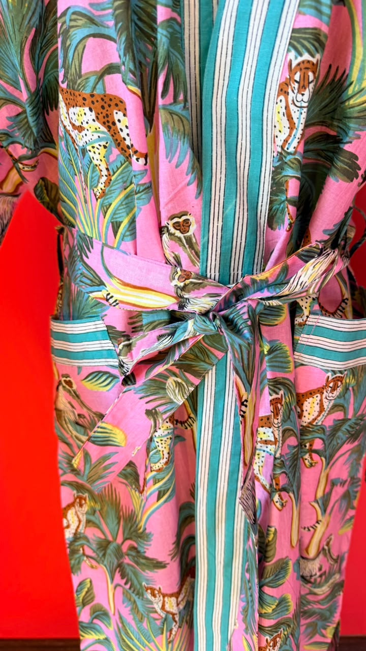 A close-up of a vibrant pink kimono robe featuring a tropical jungle print with leopards, palm leaves, and a striped tie belt, set against a solid red background.