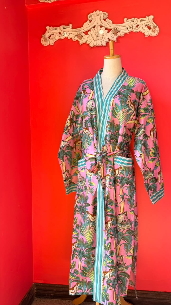 A pink tropical print robe with turquoise and white striped trim, displayed on a mannequin against a bright red wall.