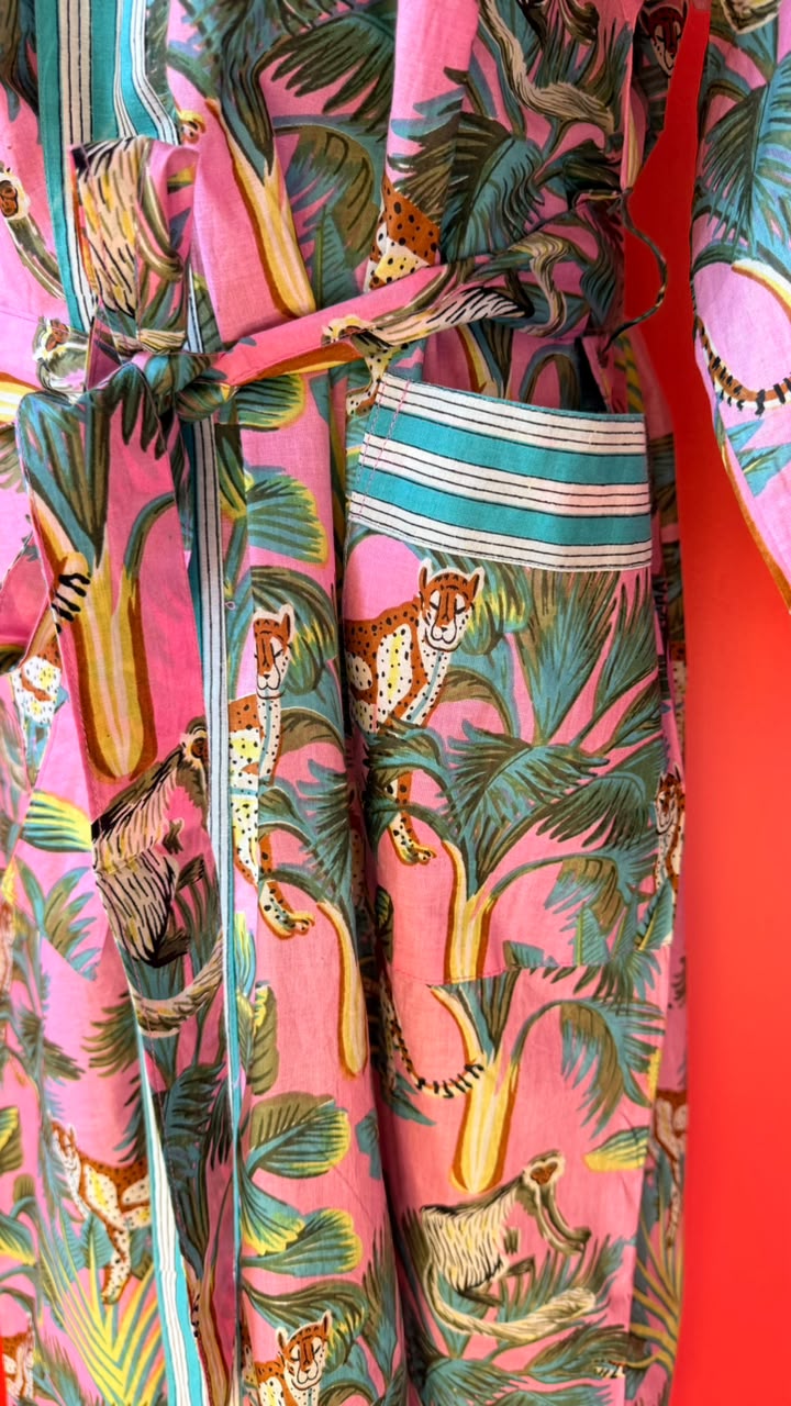 A close-up of a vibrant pink patterned garment featuring a tropical jungle print with palm trees, leopards, and a striped waist tie.