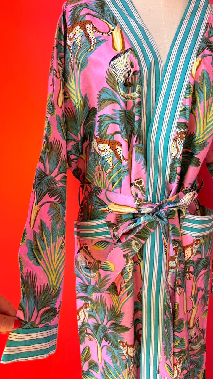 A long-sleeve wrap robe with a vibrant pink background featuring a tropical print of palm leaves, monkeys, and leopards, accented with teal and white vertical stripes on the collar, front opening, and tie belt.