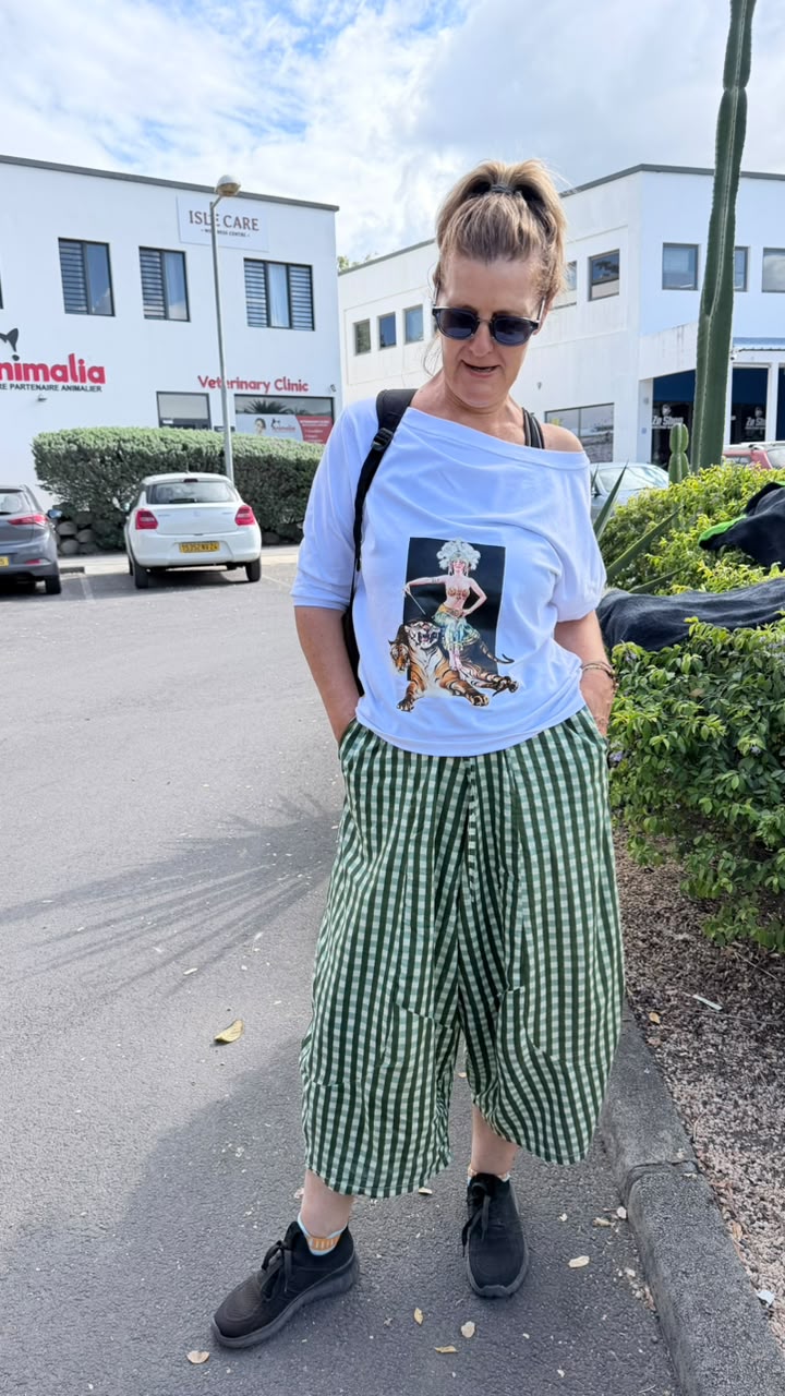 A woman standing outdoors on a street wearing a light blue off-the-shoulder graphic T-shirt and green and white gingham cropped trousers, with black sneakers and sunglasses.